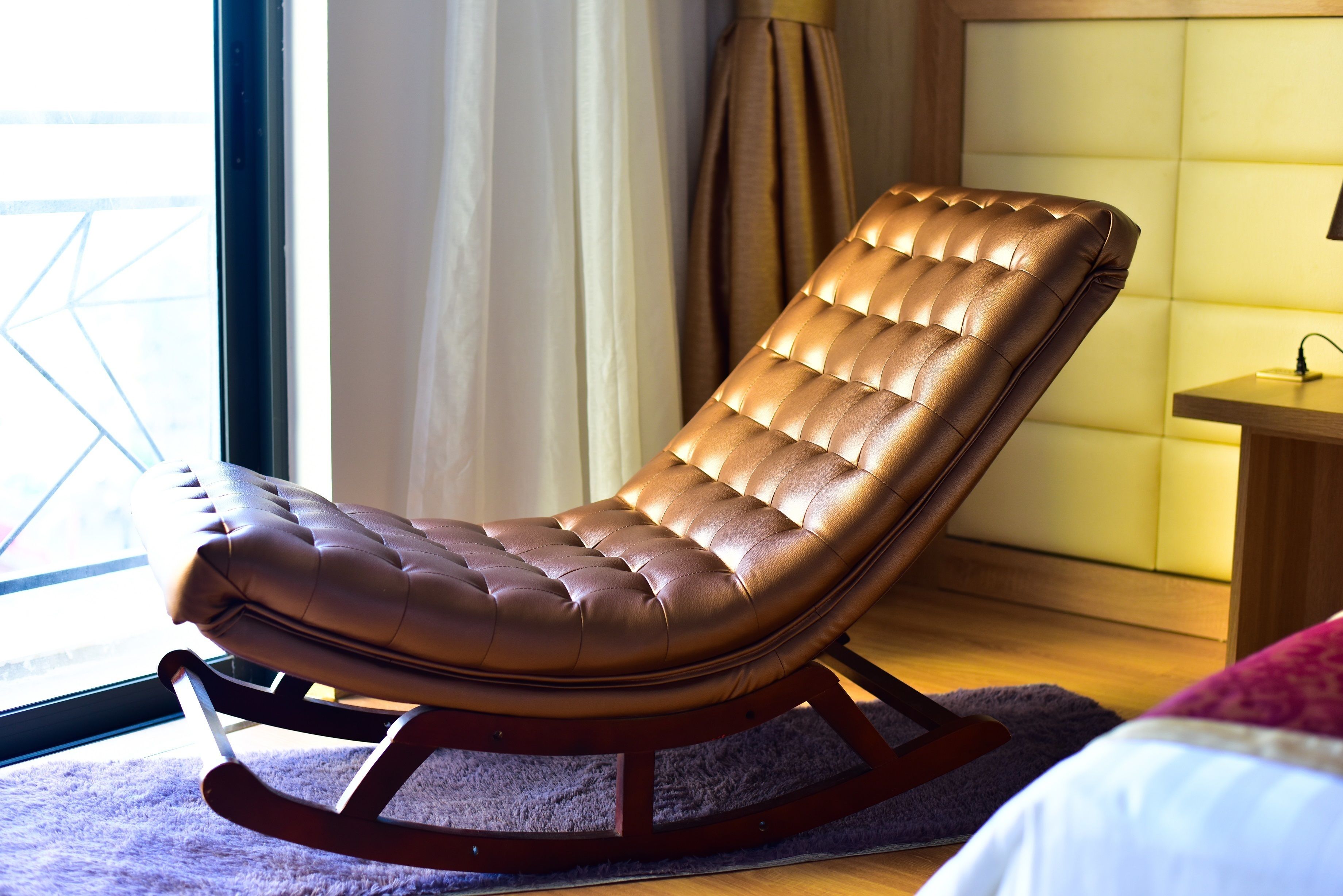 A brown leather reclining chair with a wooden base, positioned near a window with curtains, on a purple rug.