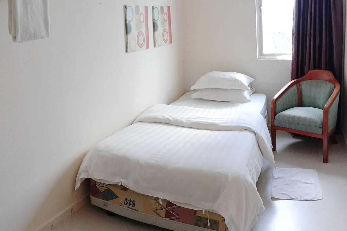 Ein kleines, schlichtes Zimmer mit einem Bett, einem Stuhl und einem Fenster mit Vorhängen.