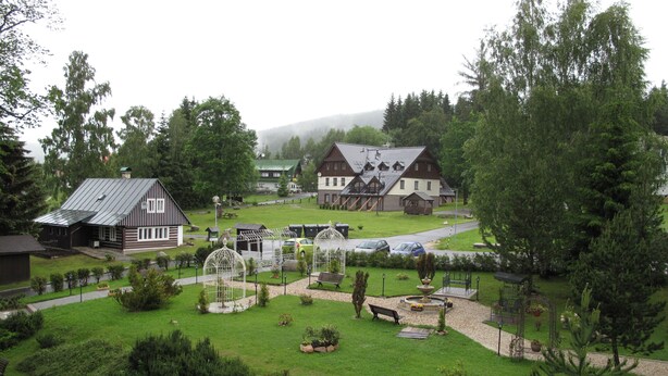 Ein ländliches Dorf mit traditionellen Holzhäusern, einem zentralen Platz mit Bänken und einem kleinen Brunnen.