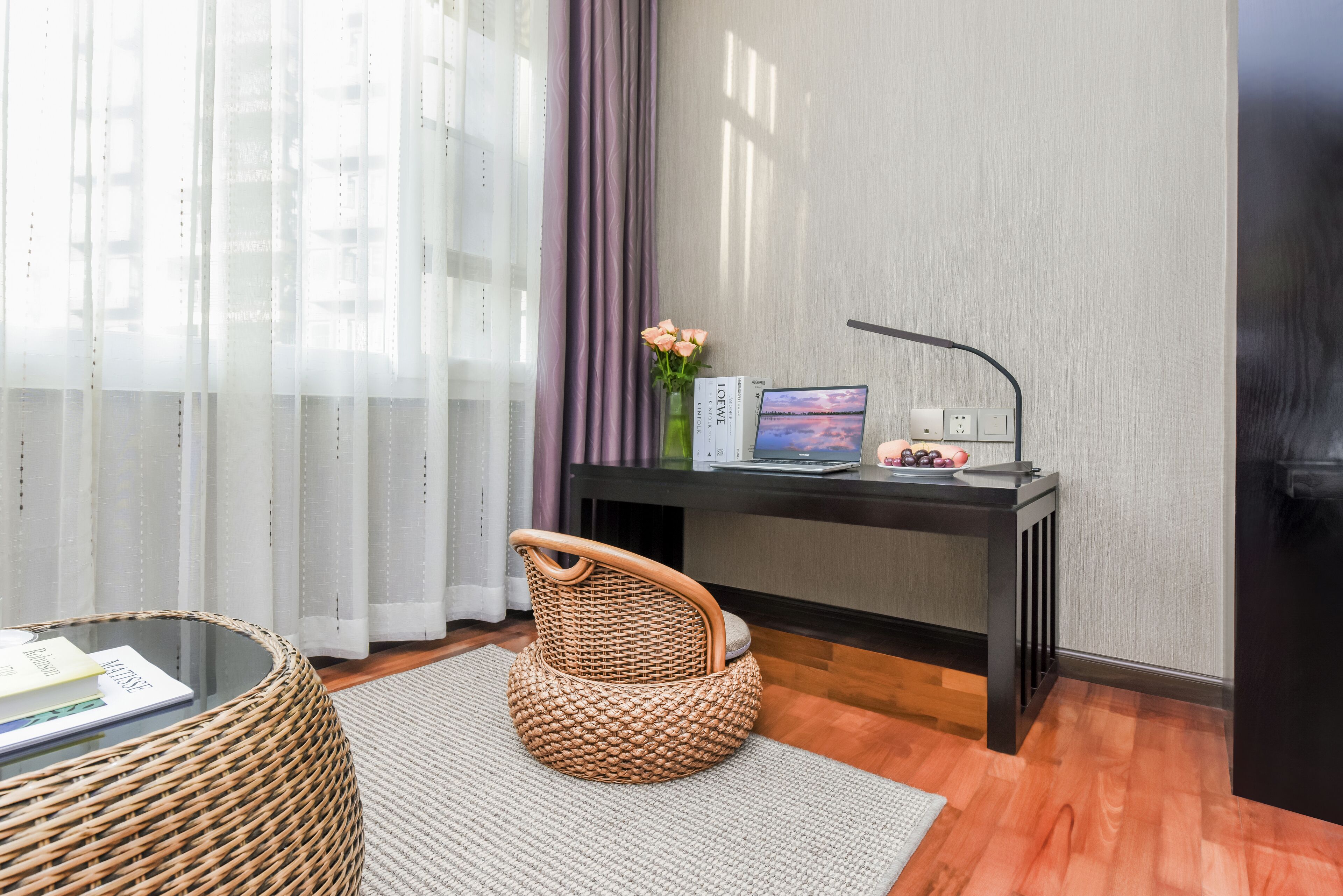 Ein modernes Zimmer mit einem Glastisch, einem Korbstuhl, einem Schreibtisch mit Computer und einer Blumenvase.