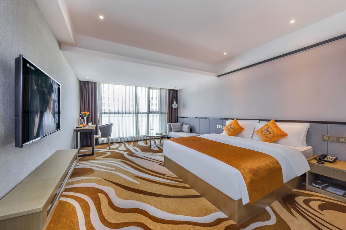 A modern hotel room with a large bed, a flat-screen TV mounted on the wall, a desk with a chair, a sofa, and a large window with curtains.