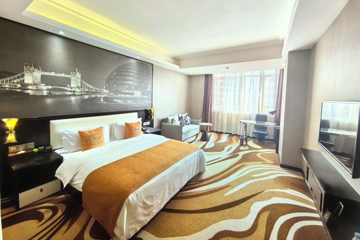 A hotel room with a large bed, a desk with a chair, a television, and a large window with curtains.
