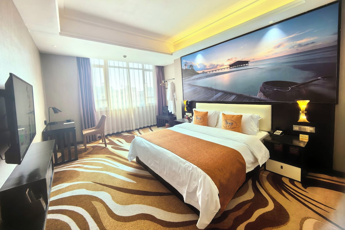 A hotel room with a large bed, a desk, a TV, and a painting of a sunset over water.