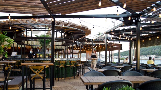 An outdoor restaurant with wooden tables and chairs, a bar area, and string lights. - Kerman Hotel (Cesme)