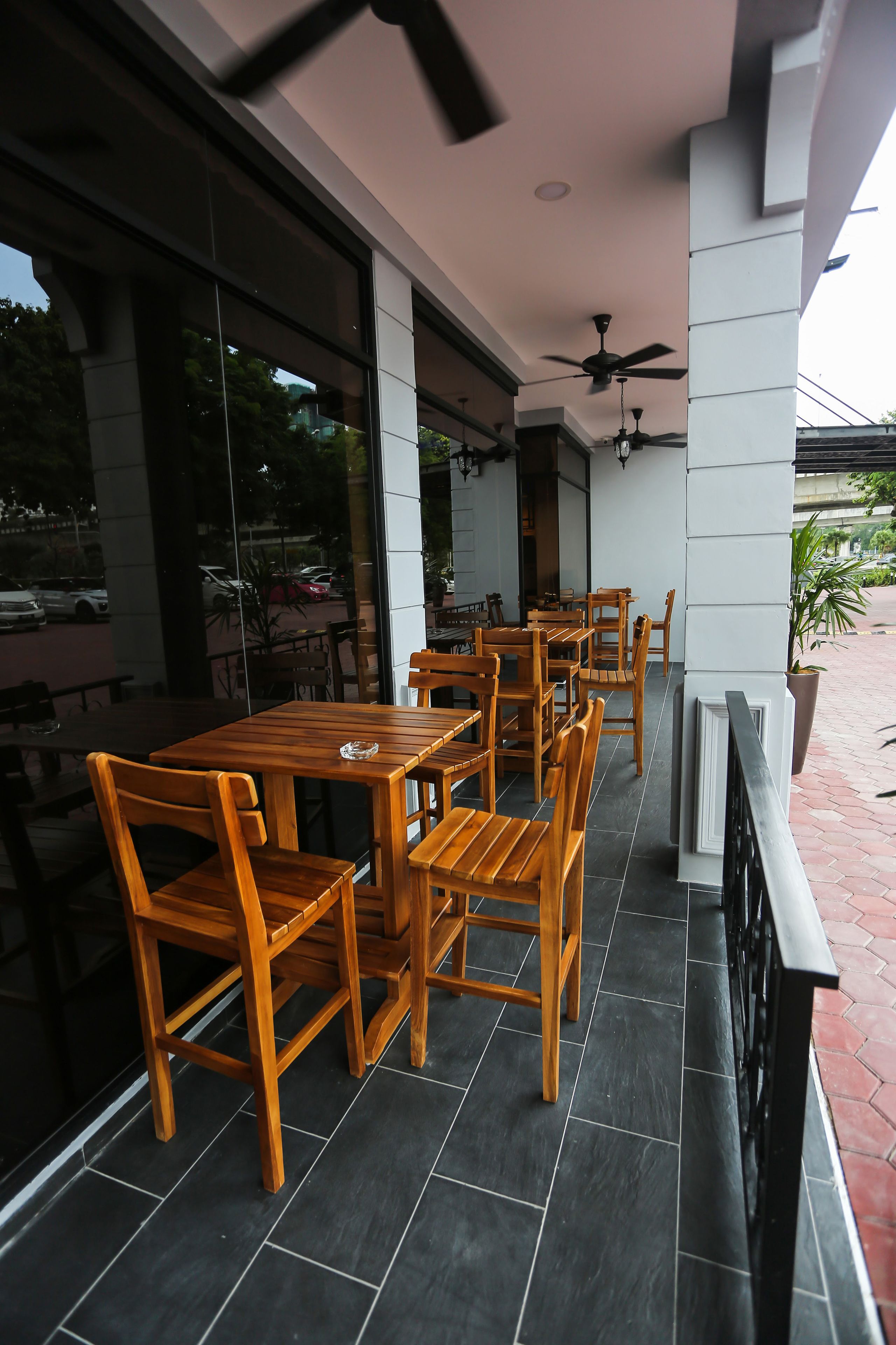 A covered outdoor seating area with wooden tables and chairs, large windows, and ceiling fans.