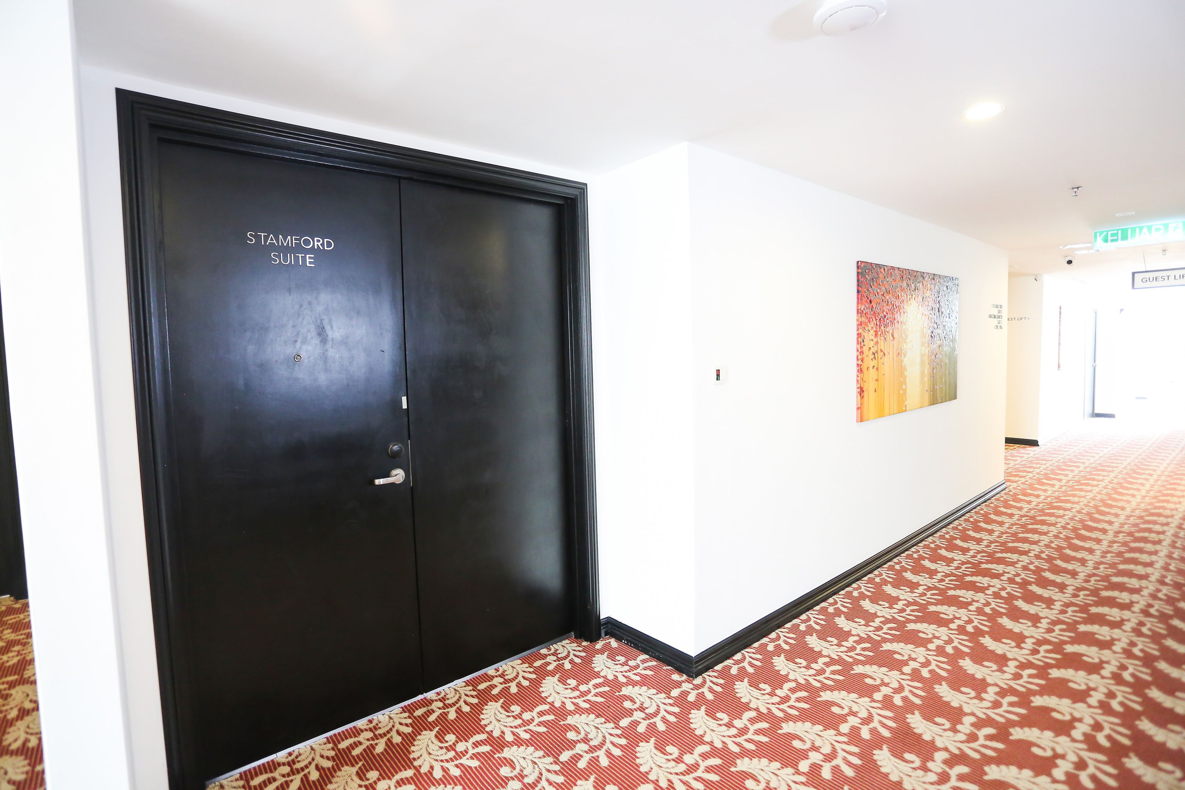 A hallway with a red patterned carpet, a black door labeled "STAMFORD SUITE", and a colorful abstract painting on the wall.