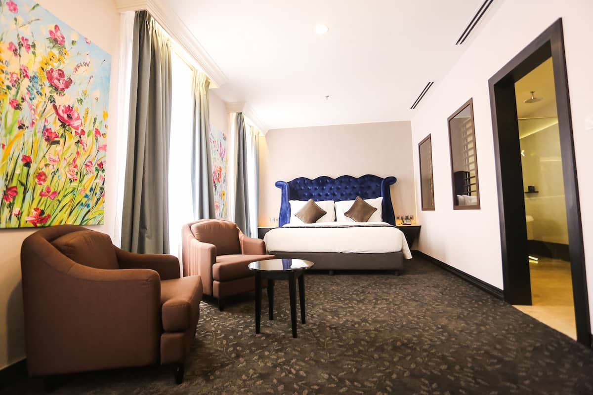 Une chambre d’hôtel avec un grand lit, un coin salon comprenant une chaise et une petite table, un tableau au mur et une porte donnant sur une autre pièce.
