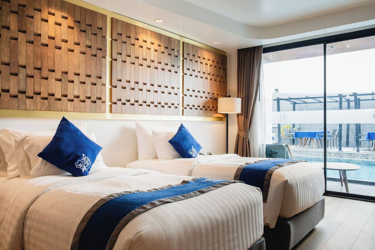 A hotel room with two beds, a wooden headboard, a large window with a view of the outdoors, and a small table by the window.