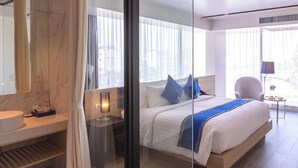 A modern hotel room with a large bed, a glass partition, a bedside table with a lamp, and a view of the city through large windows. - Wintree City Resort Chiang Mai (Chiang Mai)
