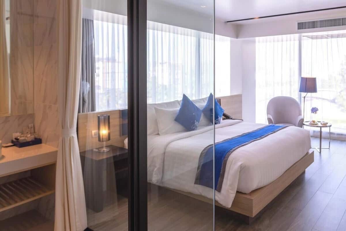 A modern hotel room with a large bed, a glass partition, a bedside table with a lamp, and a view of the city through large windows.