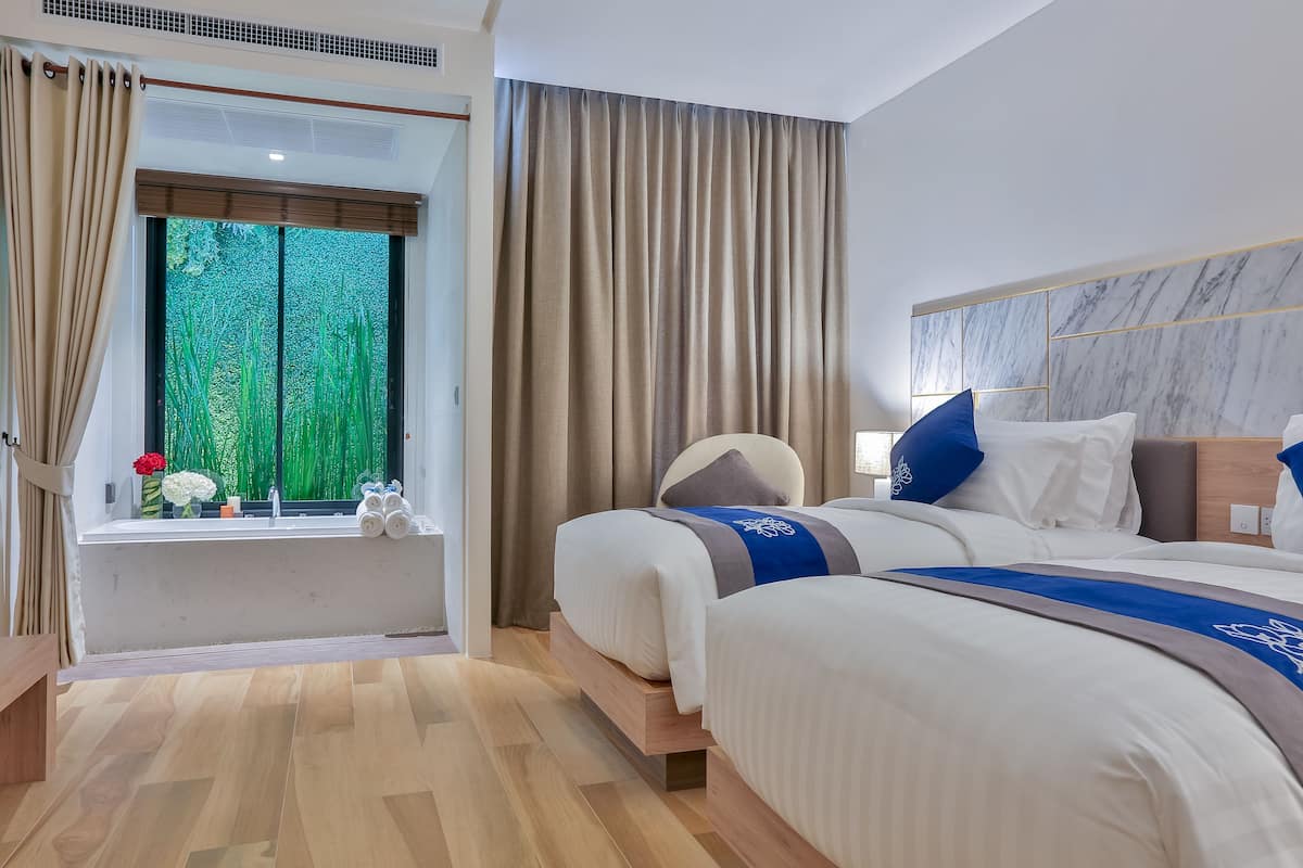 A hotel room with two beds, a large window with curtains, a marble headboard, and a view of greenery outside.
