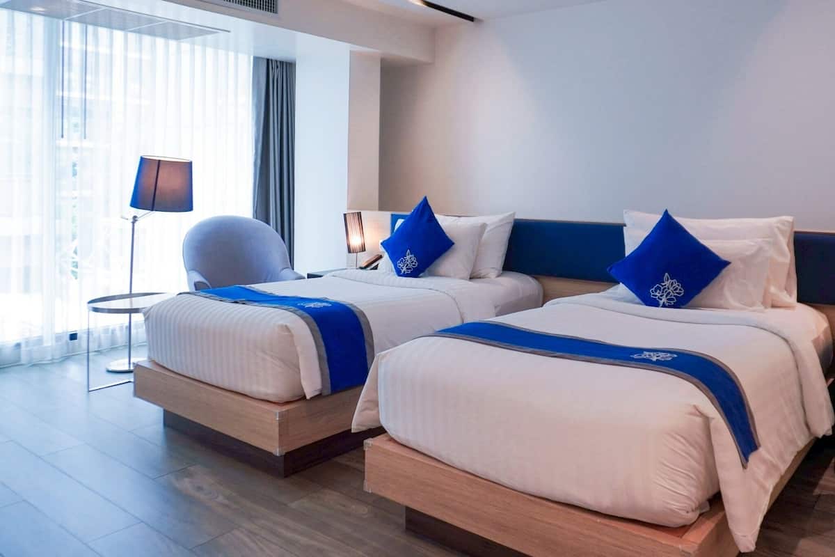A modern hotel room with two beds, a chair, a lamp, and a large window with curtains.