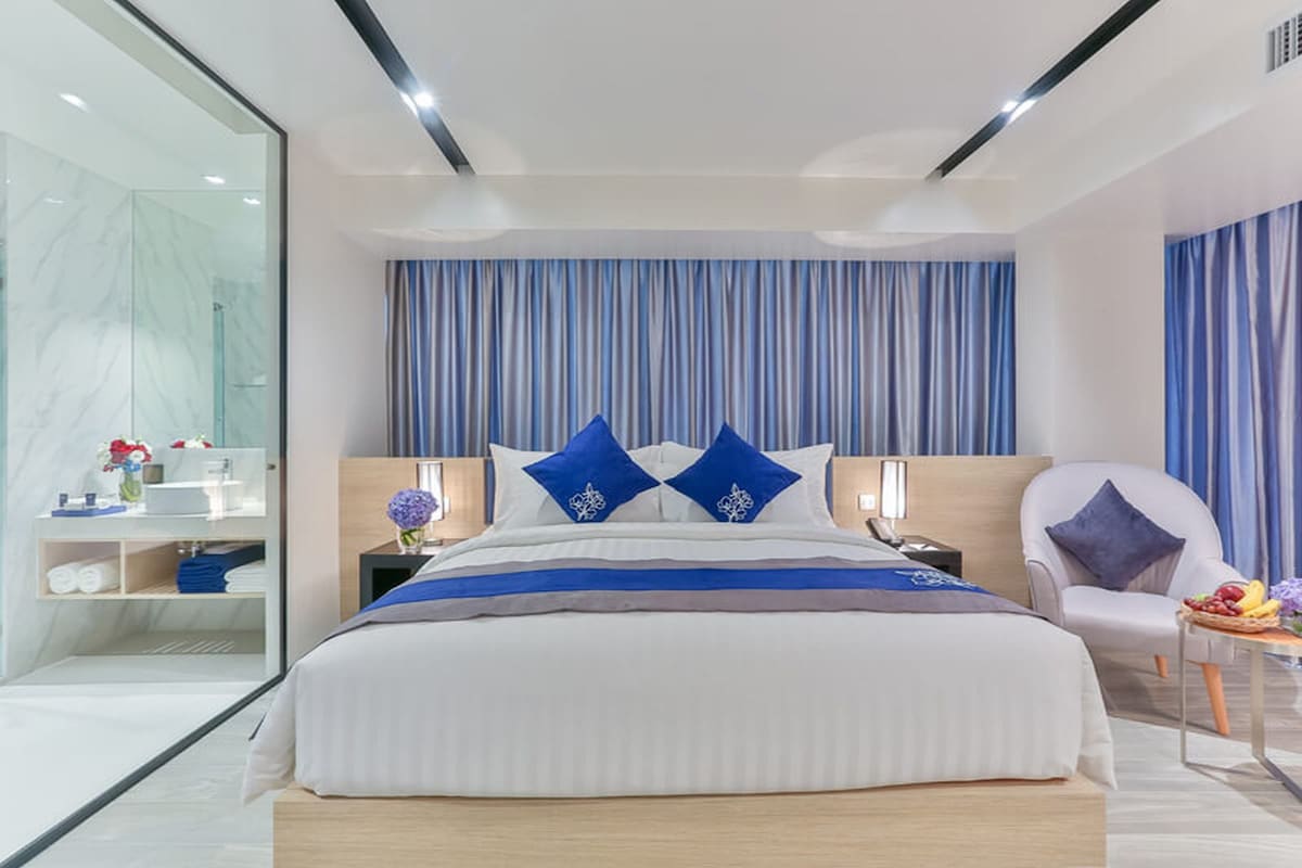 A modern hotel room with a large bed, a bedside table with a lamp, a chair, and a small table with fruit.