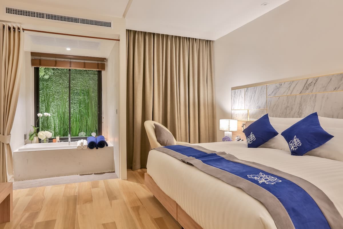A hotel room with a large bed, a window with curtains, a bedside table, and a view of greenery outside.