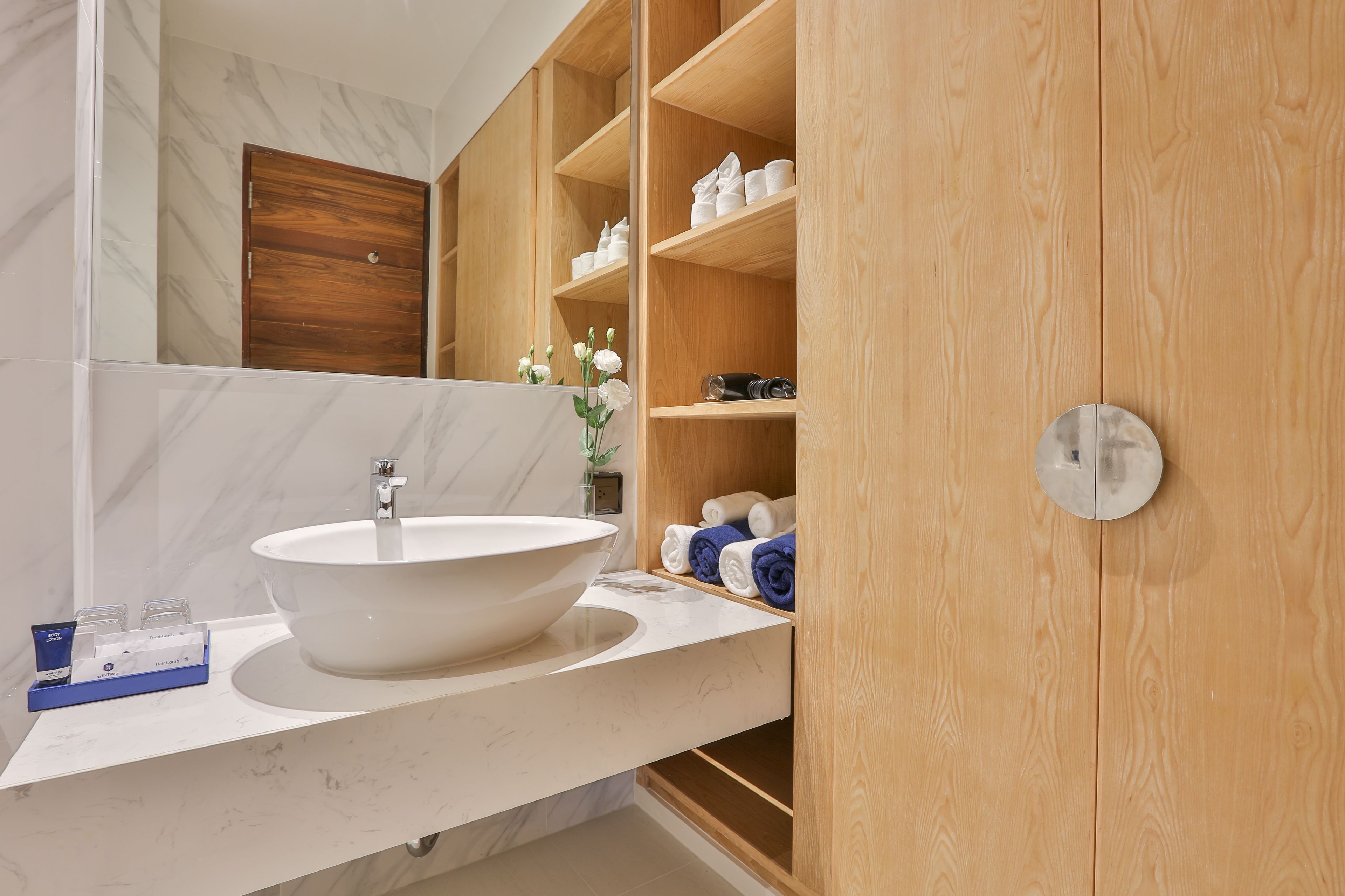 Un baño con un lavamanos grande y blanco, estantes de madera con toallas, y una pared con azulejos de mármol.