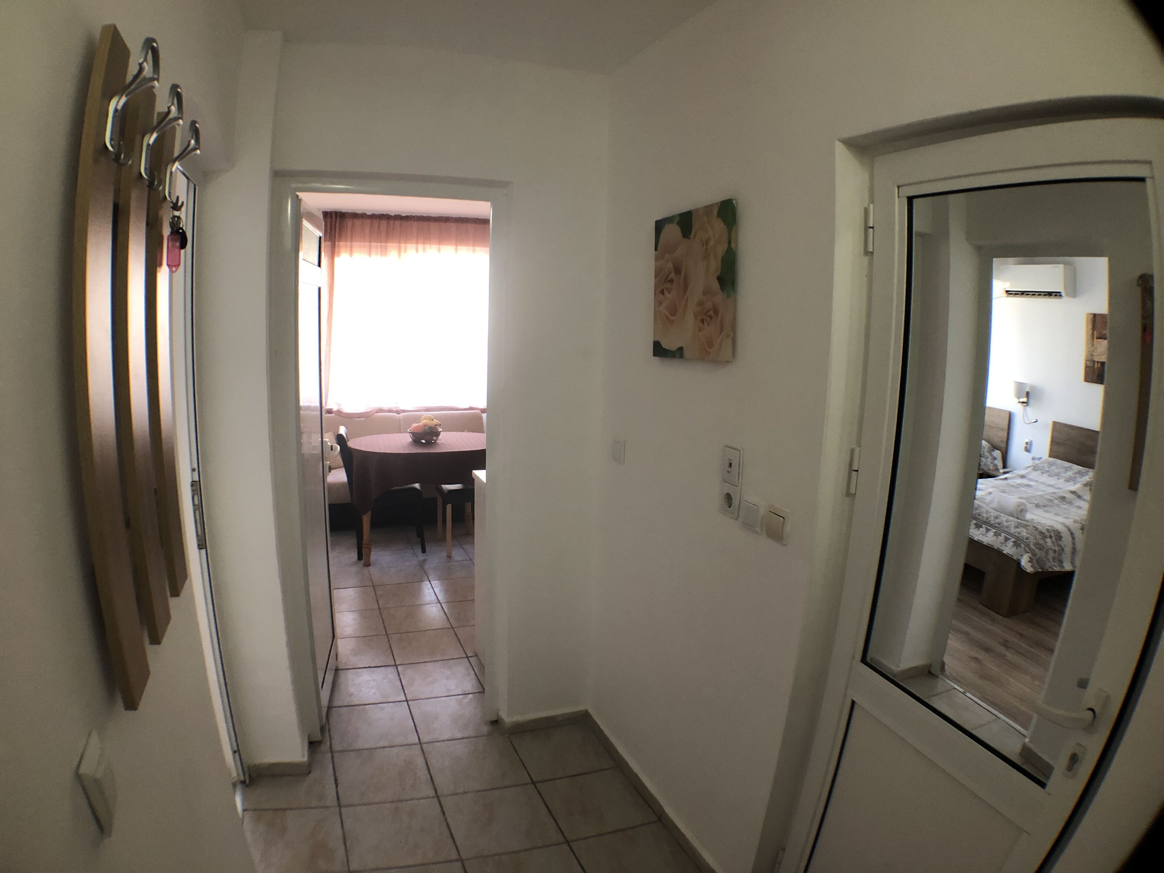 A hallway leading to a dining area with a table and chairs, a painting on the wall, and a bed visible through a door.