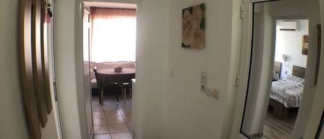 A hallway leading to a dining area with a table and chairs, a painting on the wall, and a bed visible through a door.