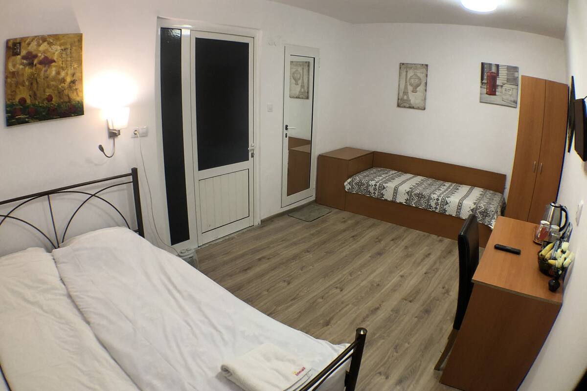 A bedroom with a bed, a sofa bed, a wooden wardrobe, a desk with a chair, and a wall-mounted TV.