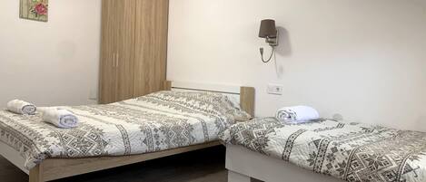 A room with two beds, a wooden wardrobe, an air conditioning unit, and a wall-mounted lamp.