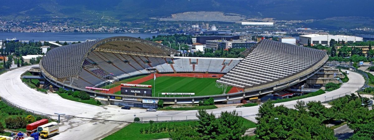 A large stadium with a curved roof, a green field, and a surrounding road.