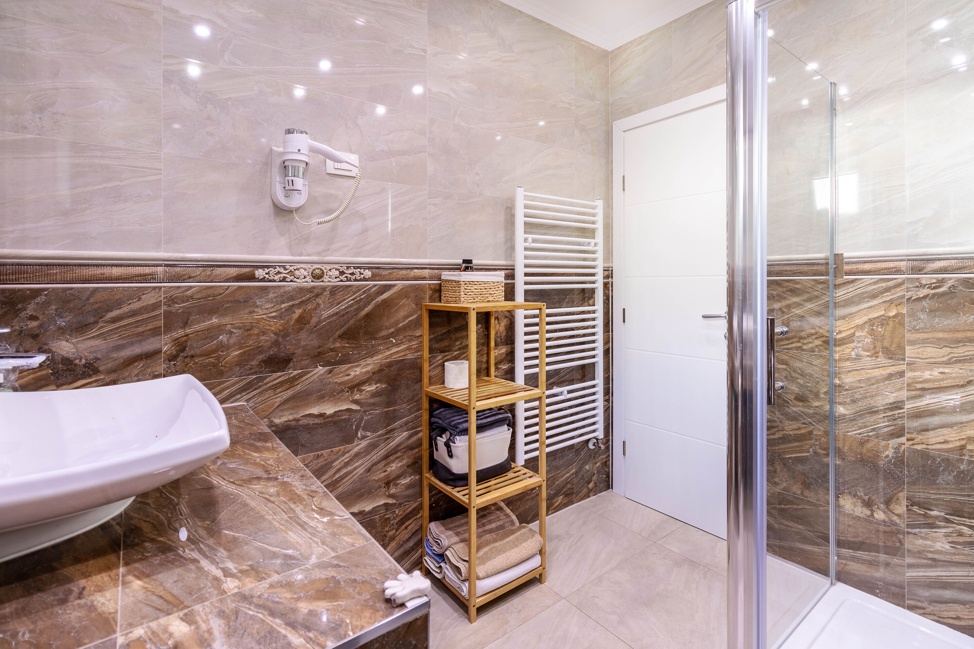 A bathroom with a marble vanity, a wooden shelf with towels, a glass shower enclosure, and a white towel rack.