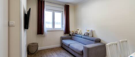 A room with a grey sofa, a window with curtains, a radiator, and a small round ottoman.