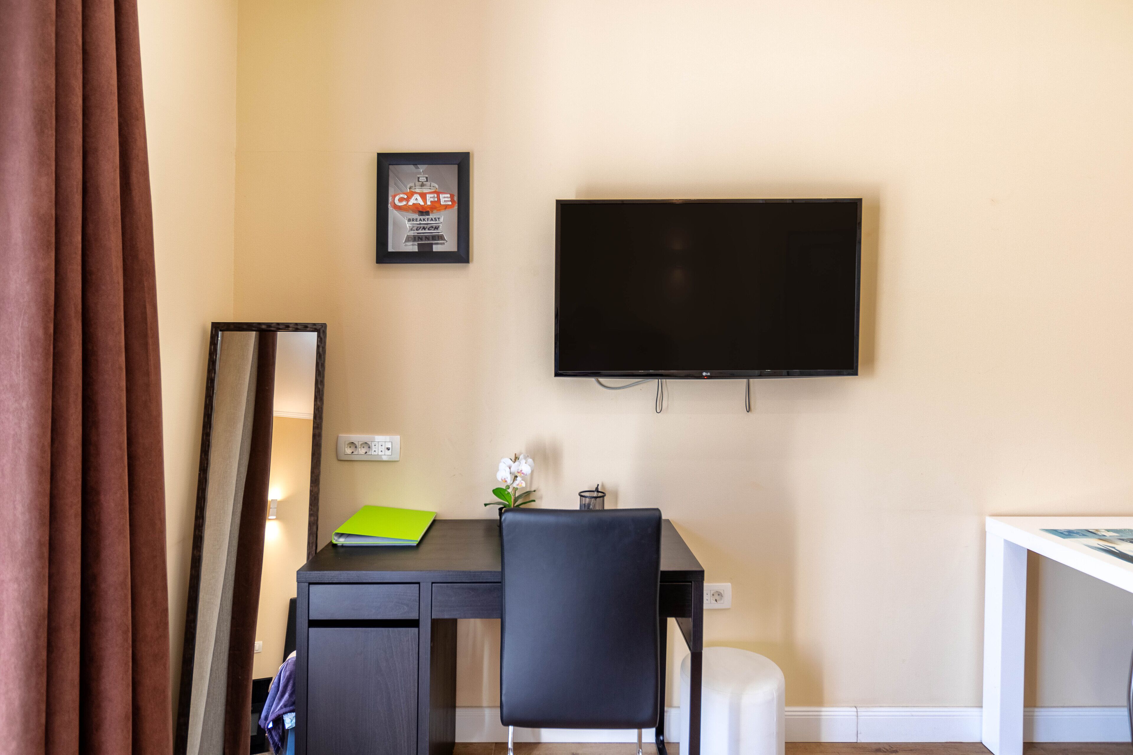 A hotel room with a desk, a chair, a TV mounted on the wall, and a framed picture of a cafe.