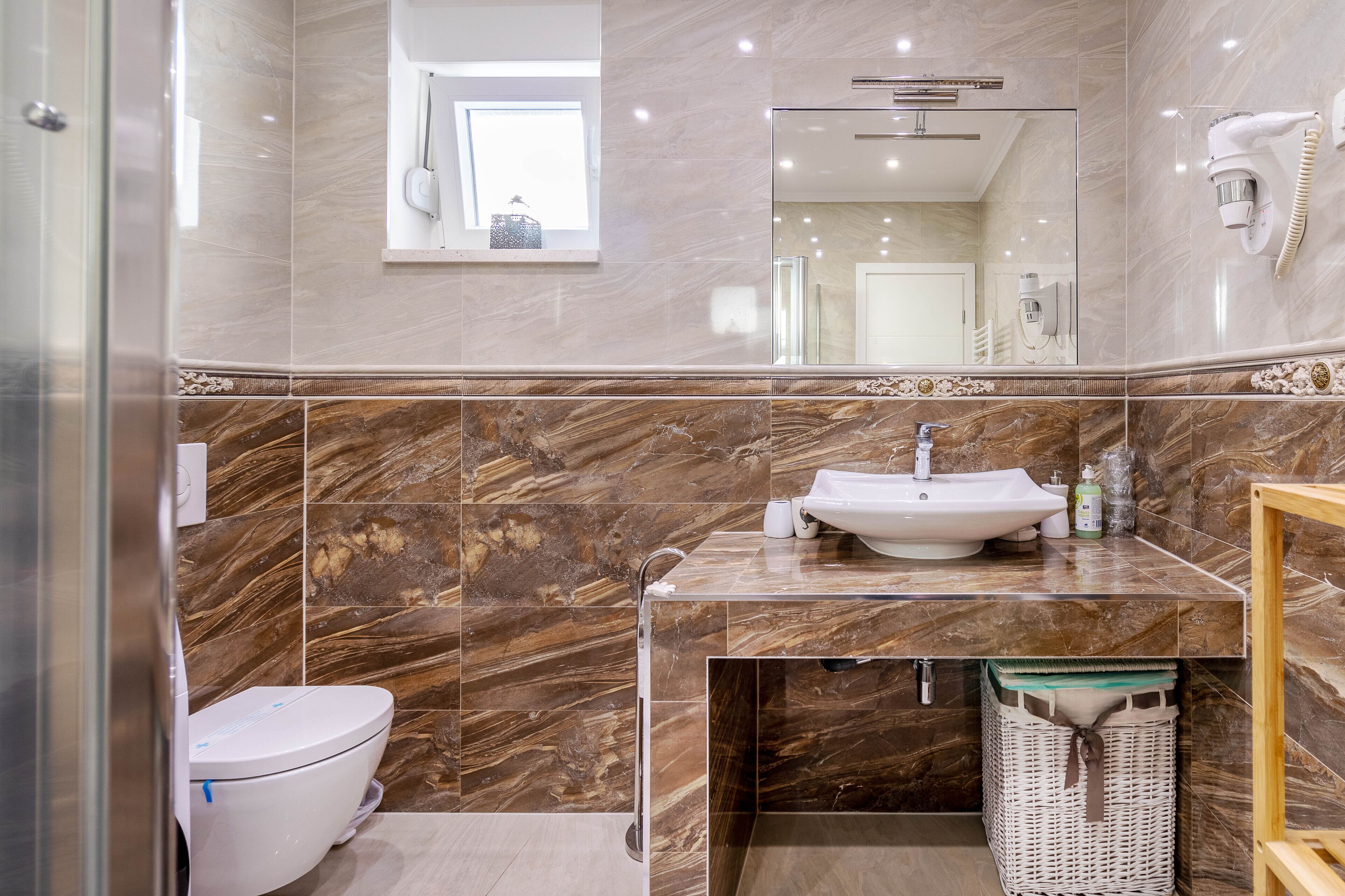A bathroom with a marble vanity, a toilet, a sink, a mirror, and a hairdryer mounted on the wall.