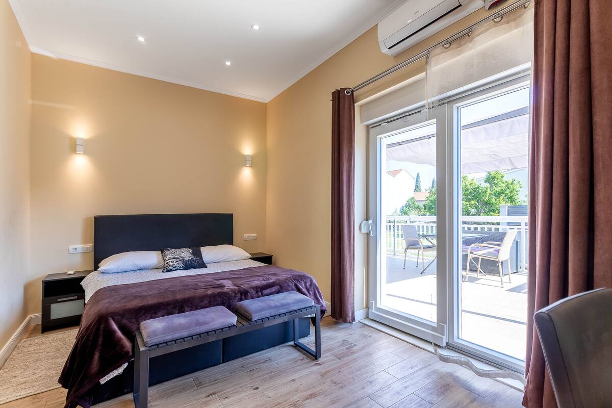 A modern bedroom with a large bed, a bench, a nightstand, and a sliding glass door leading to an outdoor area.