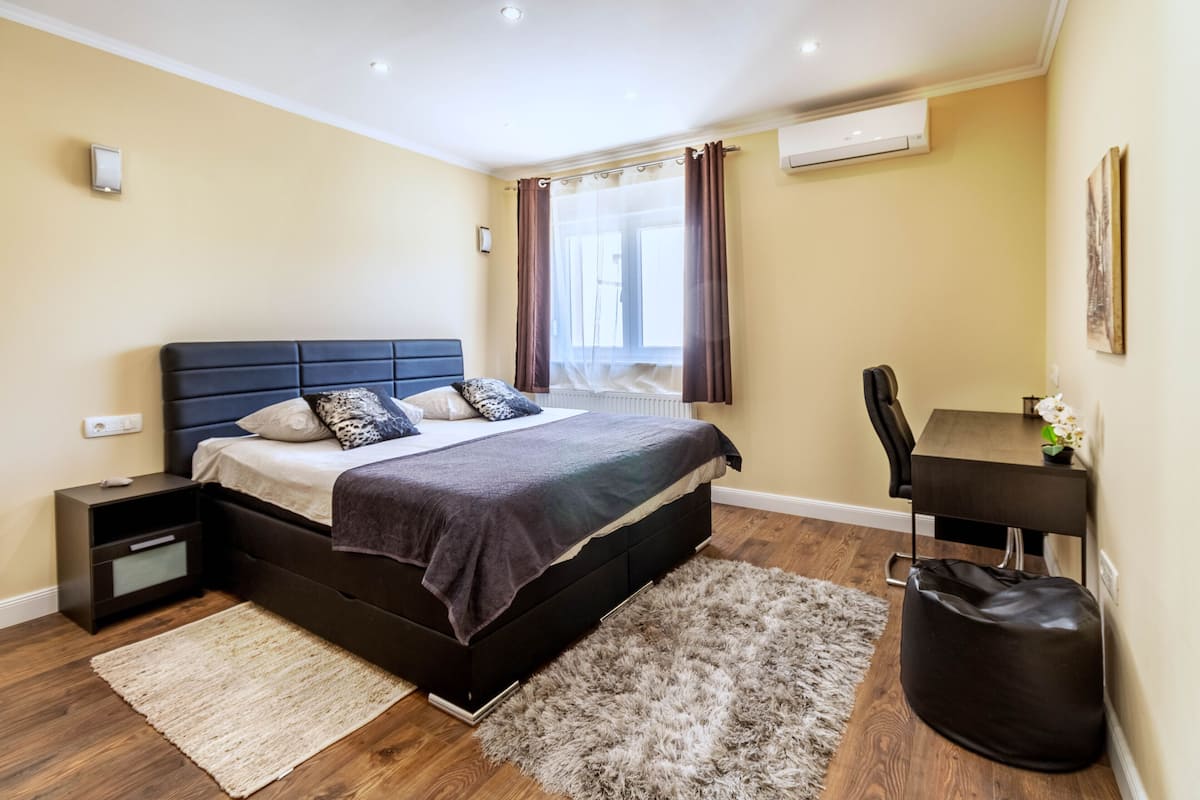 A bedroom with a bed, bedside table, desk, chair, and a bag.