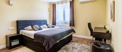 A bedroom with a bed, bedside table, desk, chair, and a bag.