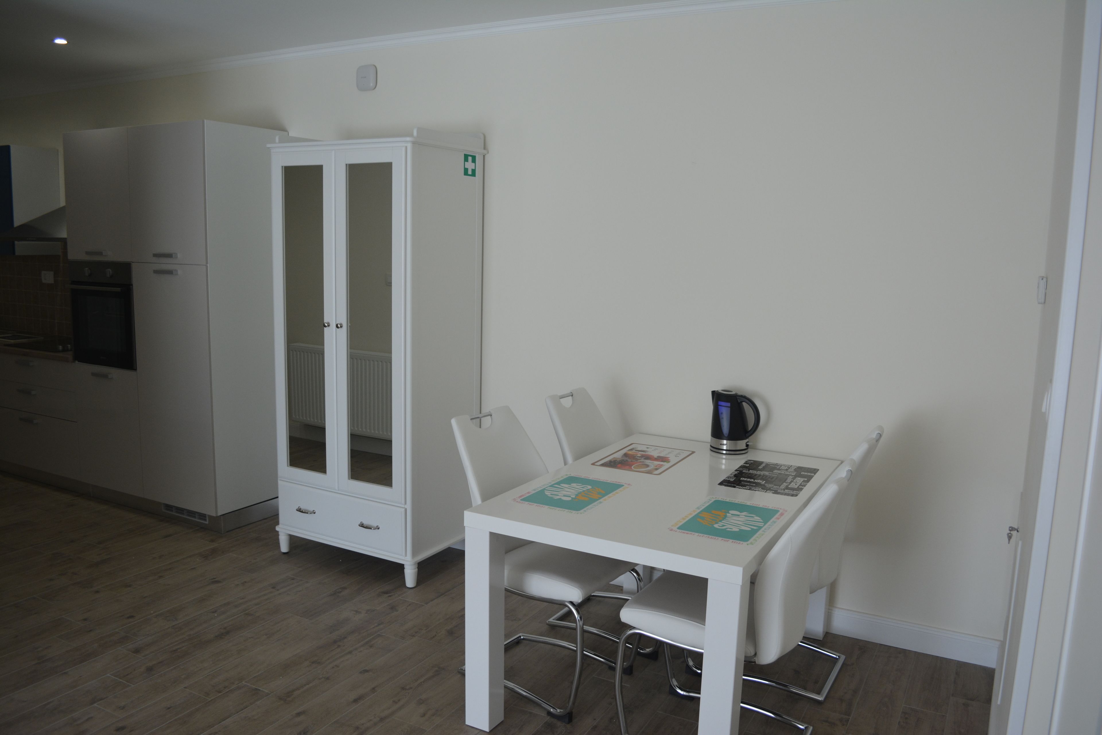 A small, clean room with a white table, four chairs, a cabinet, and a kettle.