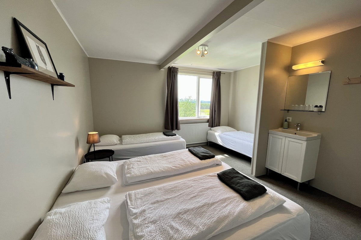 A hotel room with two beds, a bedside table with a lamp, a window with curtains, a bathroom with a sink and mirror, and a shelf with framed pictures.