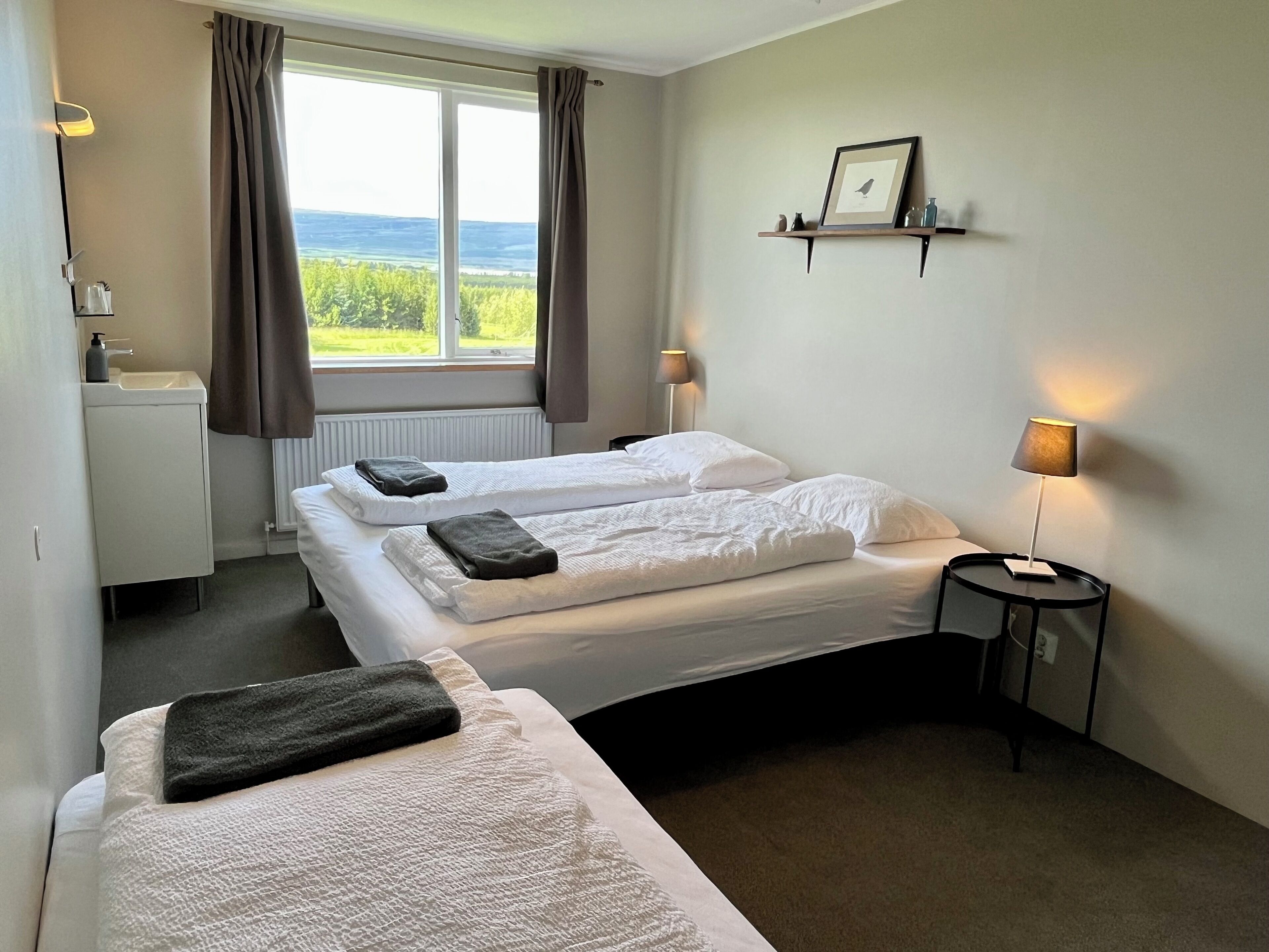 A hotel room with two beds, a small bedside table with a lamp, a window with curtains, a shelf with a picture, and a radiator.