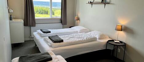 A hotel room with two beds, a small bedside table with a lamp, a window with curtains, a shelf with a picture, and a radiator.