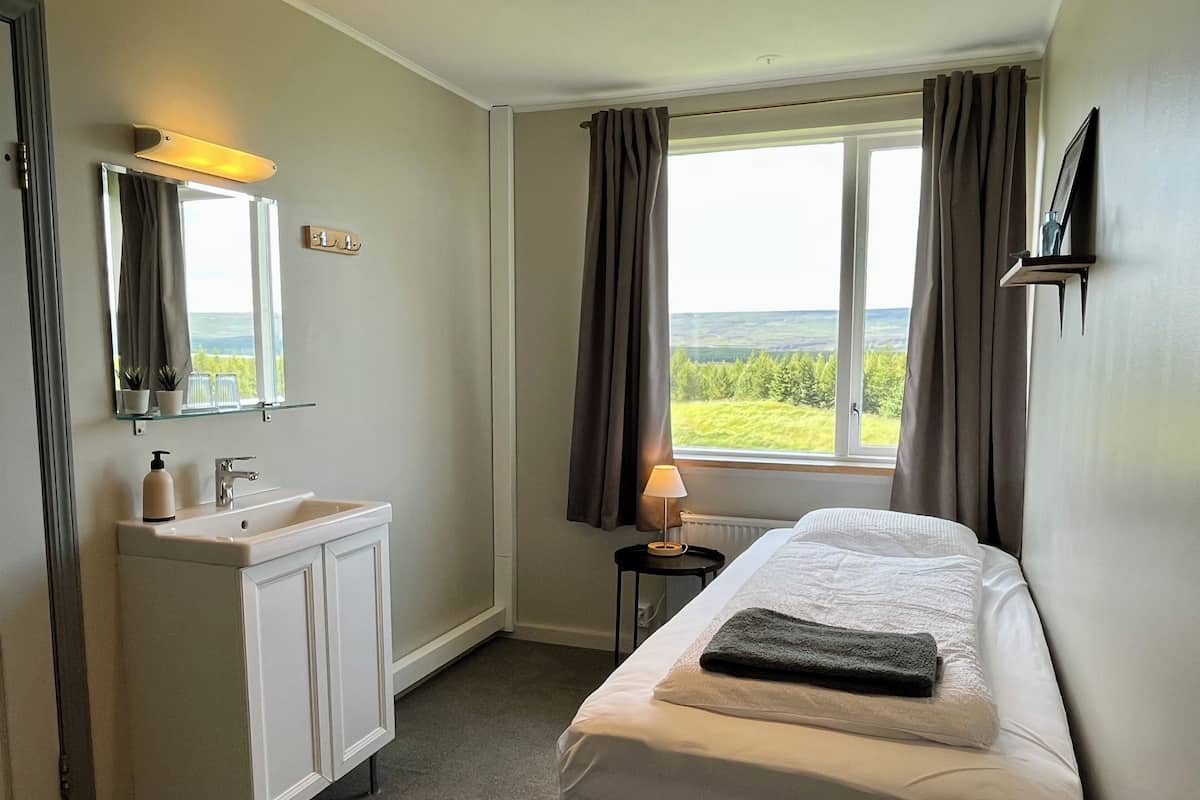 A hotel room with a bed, a bedside table with a lamp, a window with curtains, a bathroom with a sink and mirror, and a view of the outdoors.