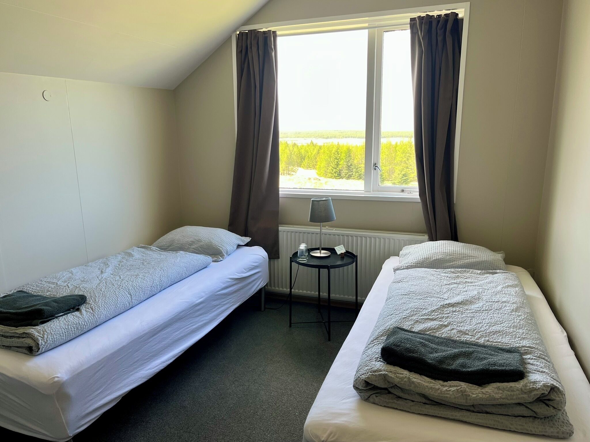 A room with two single beds, a small table with a lamp, and a window with curtains.