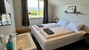 A hotel room with two beds, a window with curtains, a bedside table with a lamp, a shelf with a picture, and a sink with a soap dispenser. - Eyjólfsstaðir Guesthouse Egilsstaðir (Egilsstaðir)