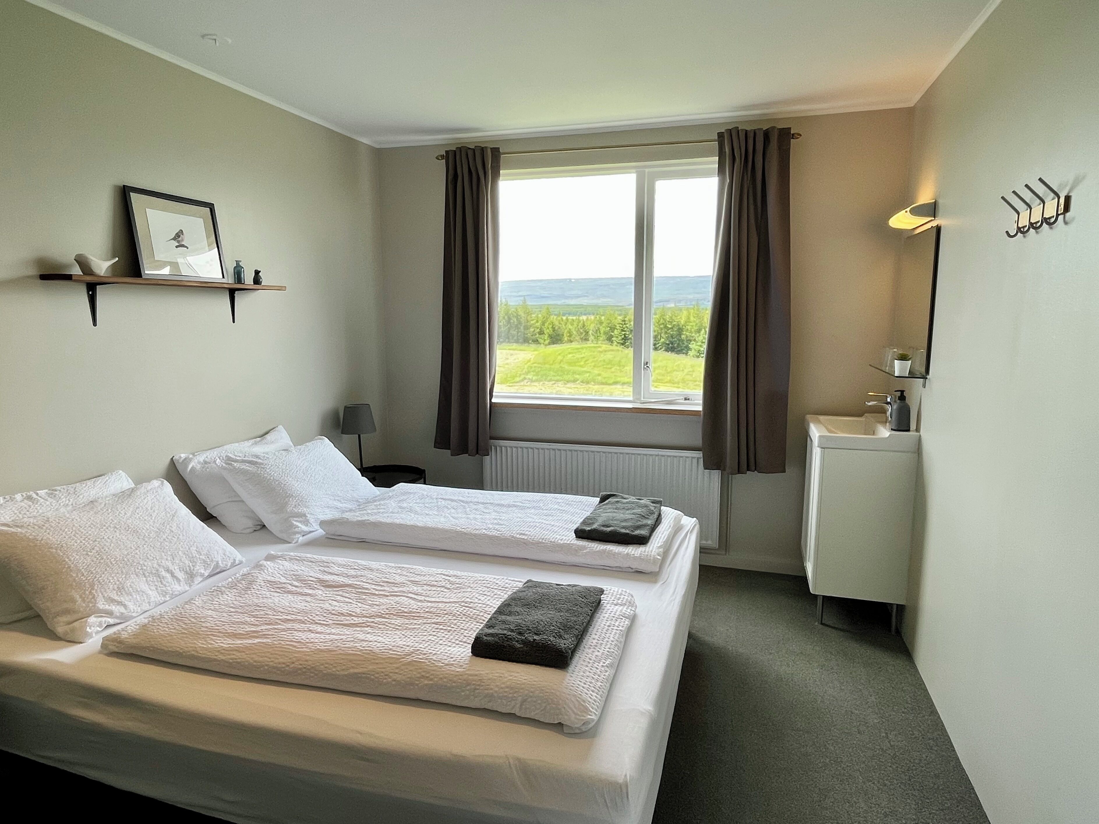 A hotel room with two beds, a window with a view of greenery, a small desk with a mirror, and a shelf with a picture.