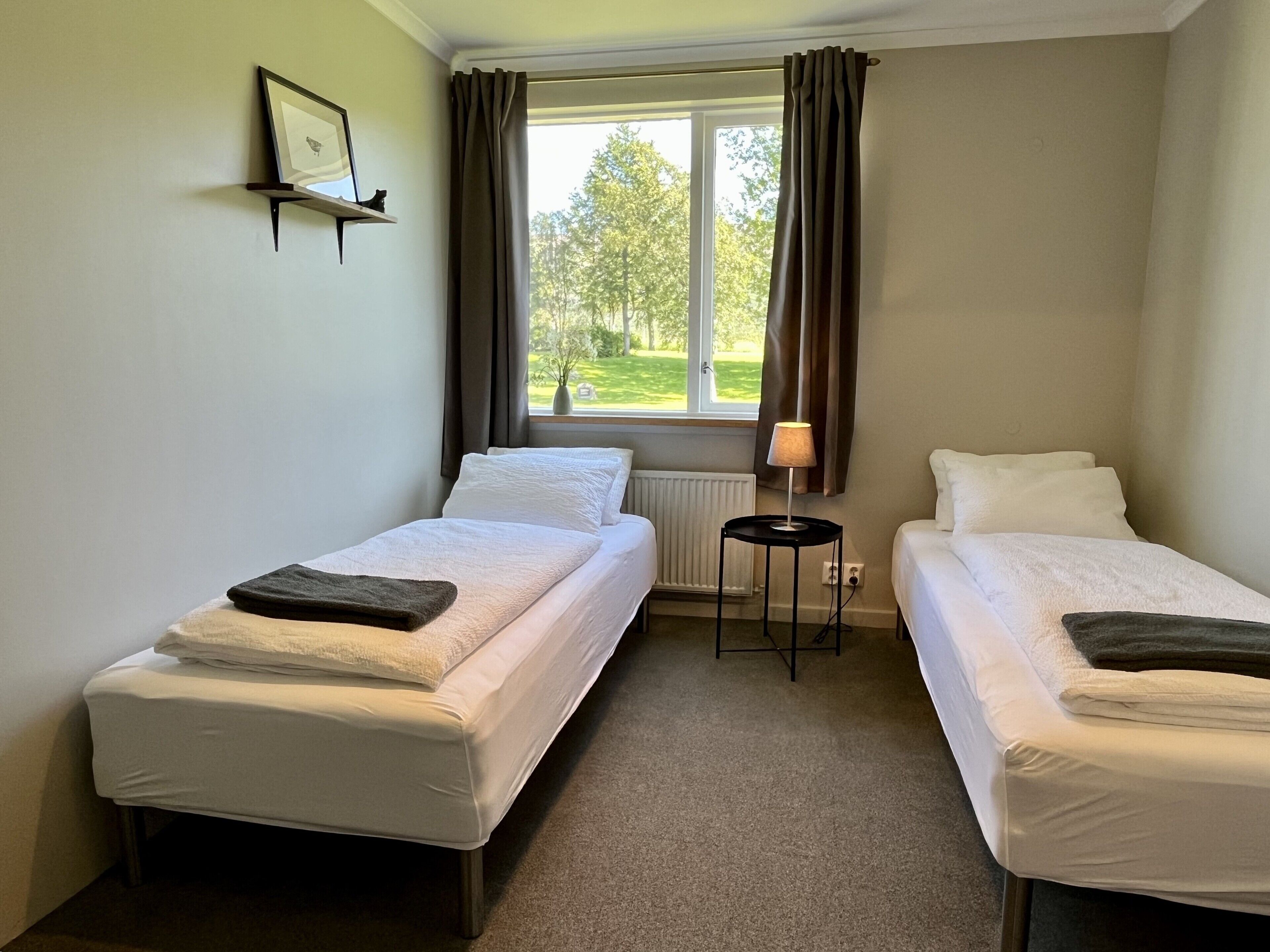 Een kamer met twee eenpersoonsbedden, een kleine tafel met een lamp, een radiator en een raam met gordijnen.
