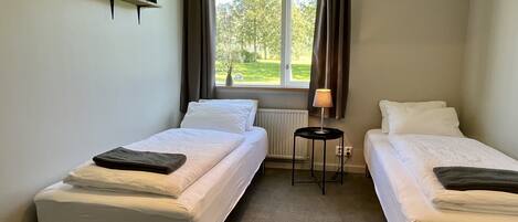 A room with two single beds, a small table with a lamp, a radiator, and a window with curtains.
