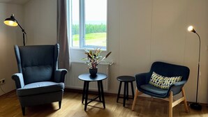 A room with a high ceiling, a large window with curtains, a blue armchair, a smaller blue sofa with a patterned cushion, a small round table with a plant, and a floor lamp. - Eyjólfsstaðir Guesthouse Egilsstaðir (Egilsstaðir)