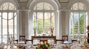 A dining area with a table set for a meal, surrounded by tall windows with a view of greenery. - Valletta Arch Apartment (Valletta)