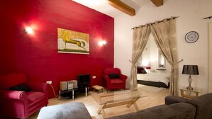 Ein Wohnzimmer mit roter Wand, einem Glastisch, einer Couch, einem roten Sessel, einem Fernseher und einem Bett mit weißem Kopfteil.