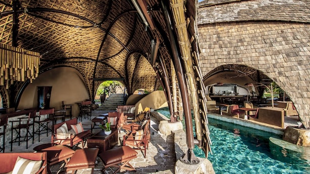 An outdoor seating area with stone tables and chairs, a pool, and a thatched roof structure.