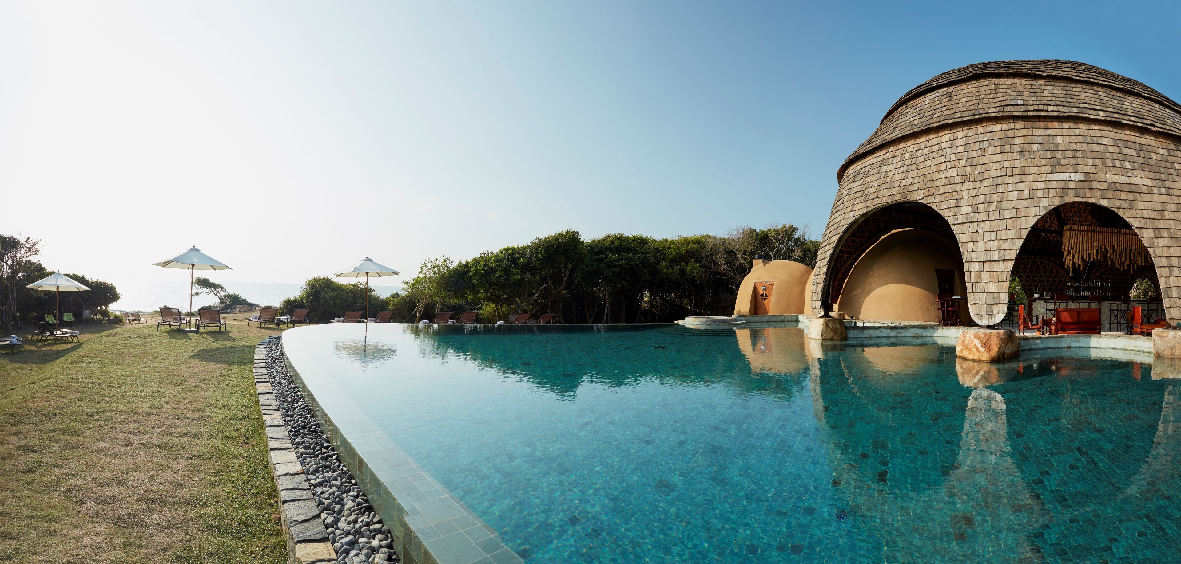 A clear swimming pool with lounge chairs and umbrellas, surrounded by a landscaped area and a distinctive building with arches.