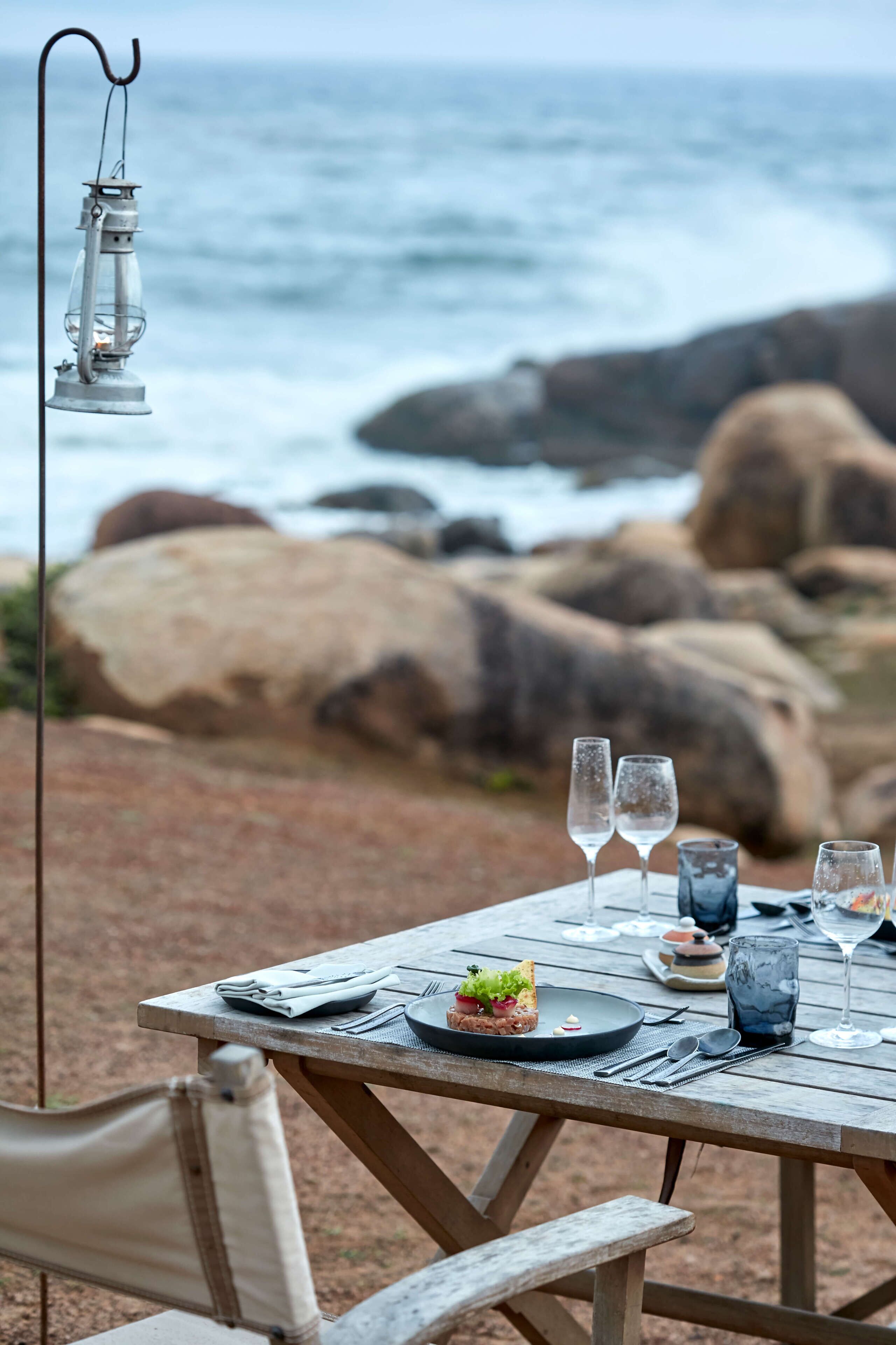 Una mesa de madera dispuesta para una comida, con vista al océano y rocas al fondo.