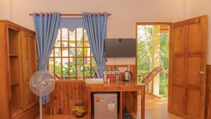 A room with wooden furniture, a small table with items, a fan, and a window with curtains. - Orchard Fruit Farm Bungalow (Phu Quoc)