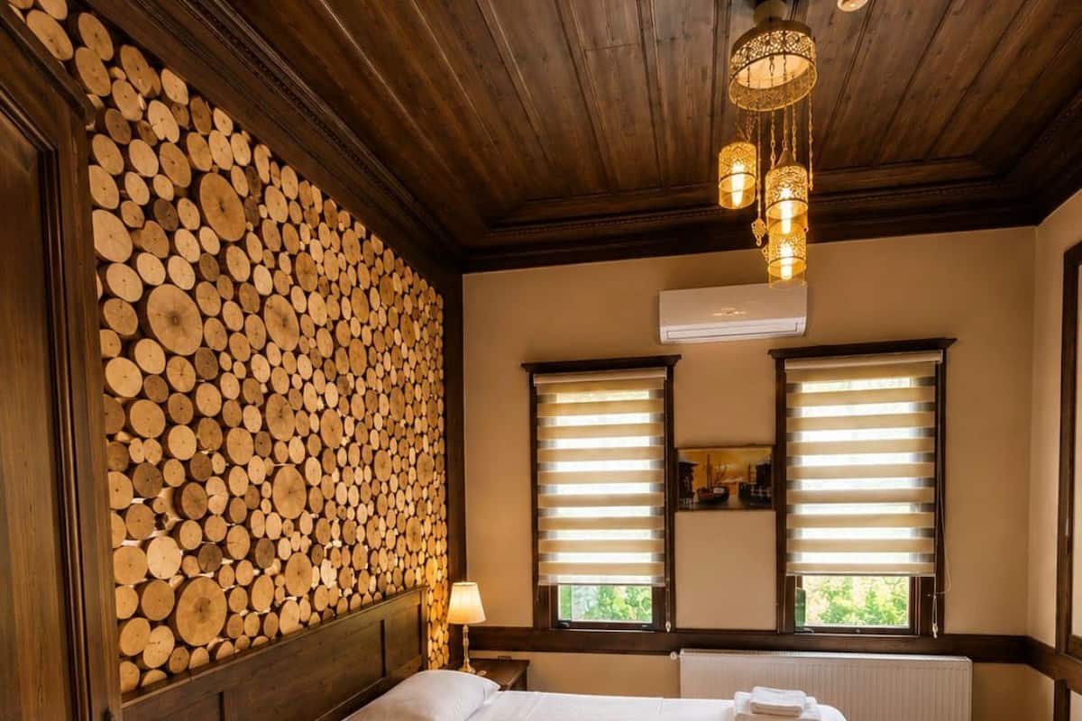 Una habitación revestida de madera con una cama, una pared apilada con troncos y dos ventanas con persianas.