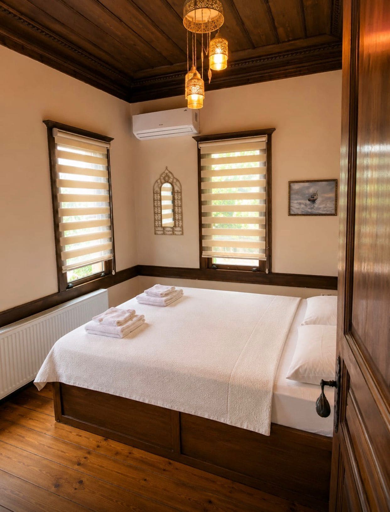 Un dormitorio con cama de madera, dos ventanas con persianas, un radiador y una lámpara colgante.